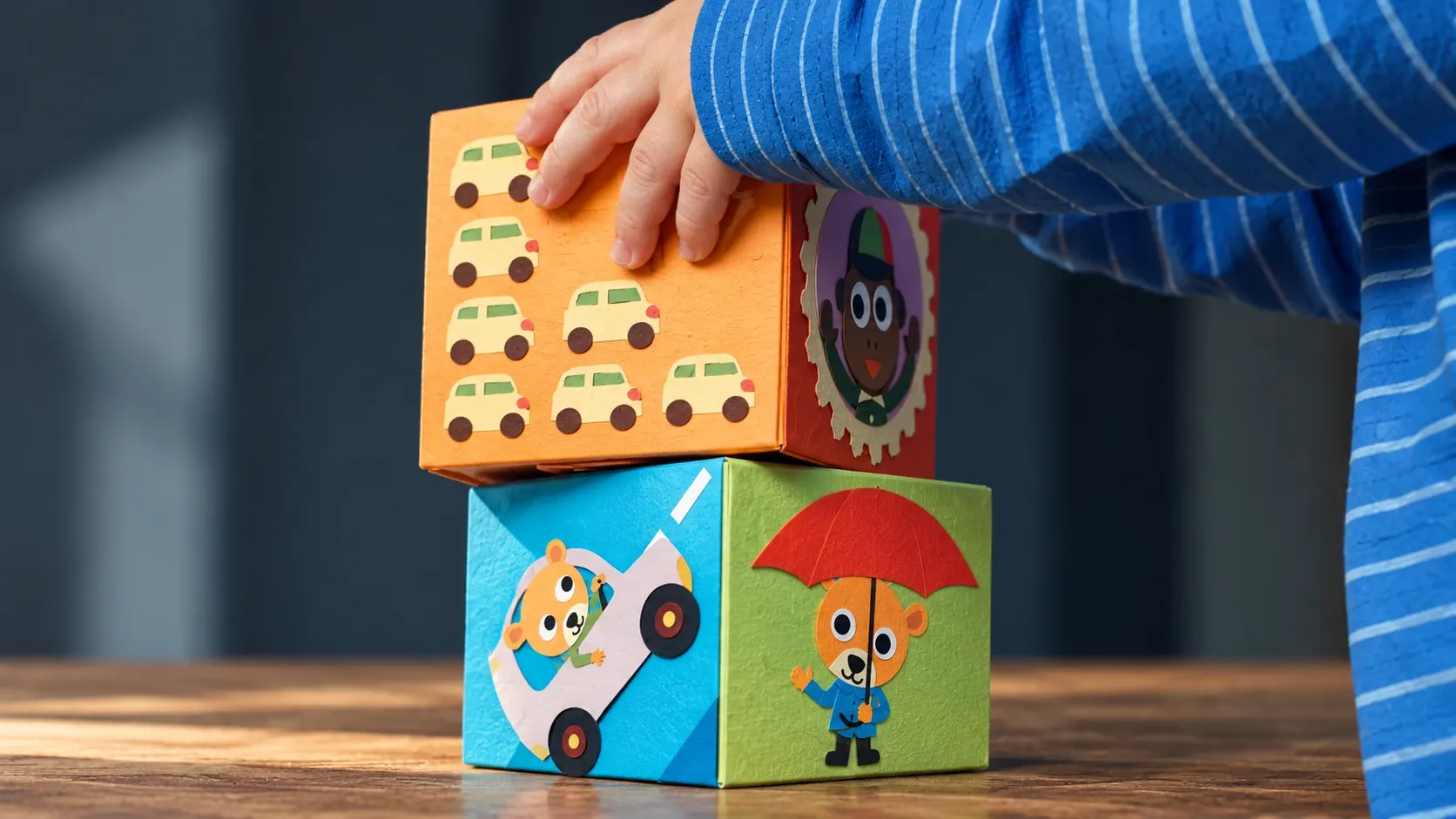 A child in a blue striped sleeve stacks colorful toy cubes decorated with paper cars and animals.